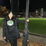 A woman standing next to a pole in front of a baseball field.