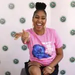 A woman in pink shirt sitting on chair with one hand up.