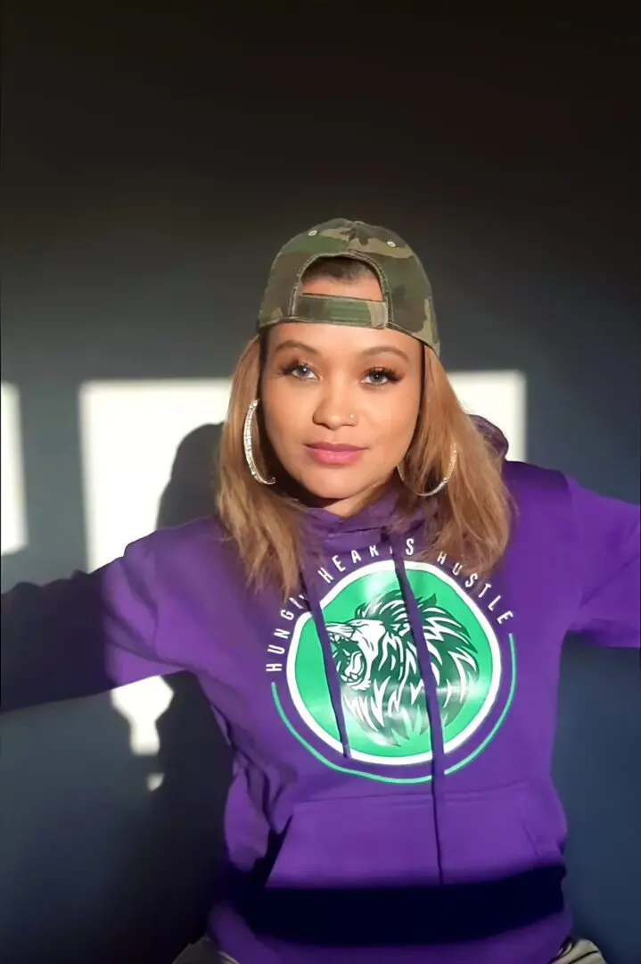 A woman in purple hoodie and hat standing next to wall.