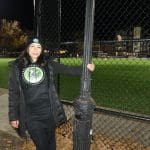 A woman standing next to a pole in front of a baseball field.